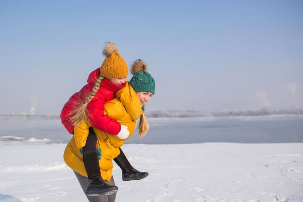 Mor bærer datteren på skuldrene i vinterlandskap, begge iført puffjakke